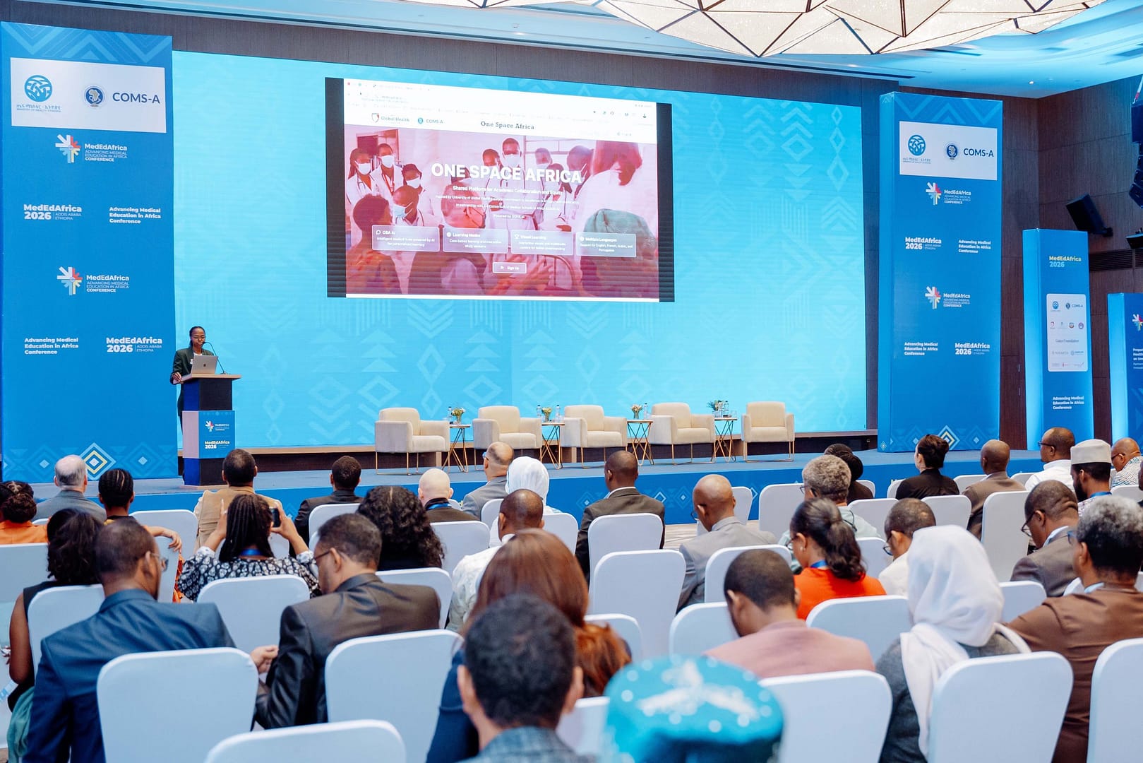 Audience seated in a large conference hall watching a speaker at a podium with a big screen showing 'ONE SPACE AFRICA' on a blue backdrop.